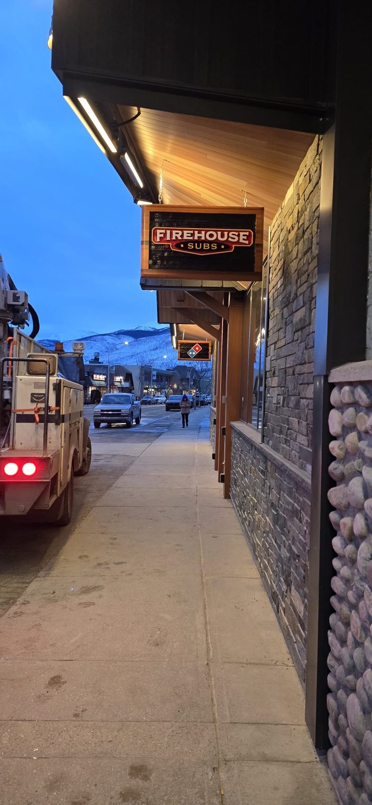 Illuminated Firehouse Subs blade sign projecting from a stone storefront at dusk