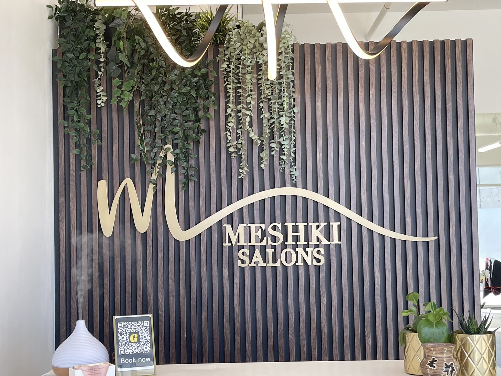 MESHKI Salons gold dimensional reception logo on a dark wood-slat feature wall